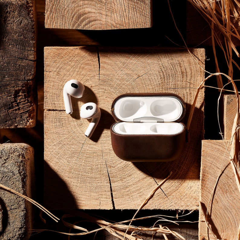 May include: A brown leather case with a white interior for wireless earbuds. The case is open and the earbuds are visible. The case is sitting on a wooden surface.
