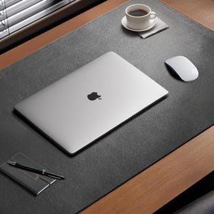 May include: A silver laptop with a black Apple logo sits on a large rectangular black leather desk mat. Beside the laptop is a white computer mouse, a black pen on a small black case, and a white coffee cup on a matching saucer.