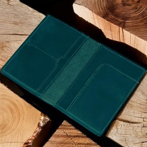 Pu&ograve; includere: Un portafoglio in pelle color teal, aperto per rivelare fessure per carte e una tasca centrale. Il portafoglio &egrave; esposto su una superficie di legno rustico, con venature visibili e toni naturali. Le cuciture sono di un teal pi&ugrave; chiaro.