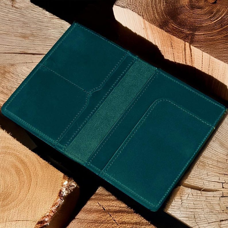 Pu&ograve; includere: Un portafoglio in pelle color teal, aperto per rivelare fessure per carte e una tasca centrale. Il portafoglio &egrave; esposto su una superficie di legno rustico, con venature visibili e toni naturali. Le cuciture sono di un teal pi&ugrave; chiaro.
