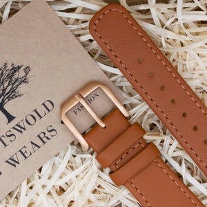 Pode incluir: Close-up de uma pulseira de relógio de couro castanho com fivela dourada. A pulseira é costurada com linha branca e tem vários orifícios. Uma etiqueta com o texto "COTSWOLD WEARS" é visível.