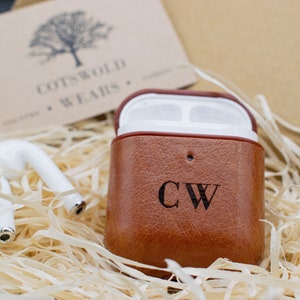 May include: A brown leather case for wireless earbuds with the initials "CW" engraved on the front. The case is sitting in a box filled with shredded paper.