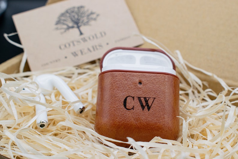 May include: A brown leather case for wireless earbuds with the initials "CW" engraved on the front. The case is sitting in a box filled with shredded paper.