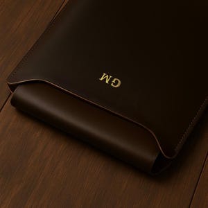 Puede incluir: Una funda de cuero marrón oscuro con las iniciales doradas "GM" grabadas en la parte delantera. La funda está abierta en la parte superior, revelando un interior marrón más claro. El artículo está colocado sobre una superficie de madera marrón oscuro.