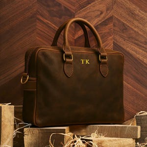 Puede incluir: Un maletín de cuero marrón con dos asas superiores y cierre de cremallera. La bolsa presenta herrajes dorados y las iniciales "TK" en dorado. La bolsa está sobre bloques de madera contra un fondo de madera.