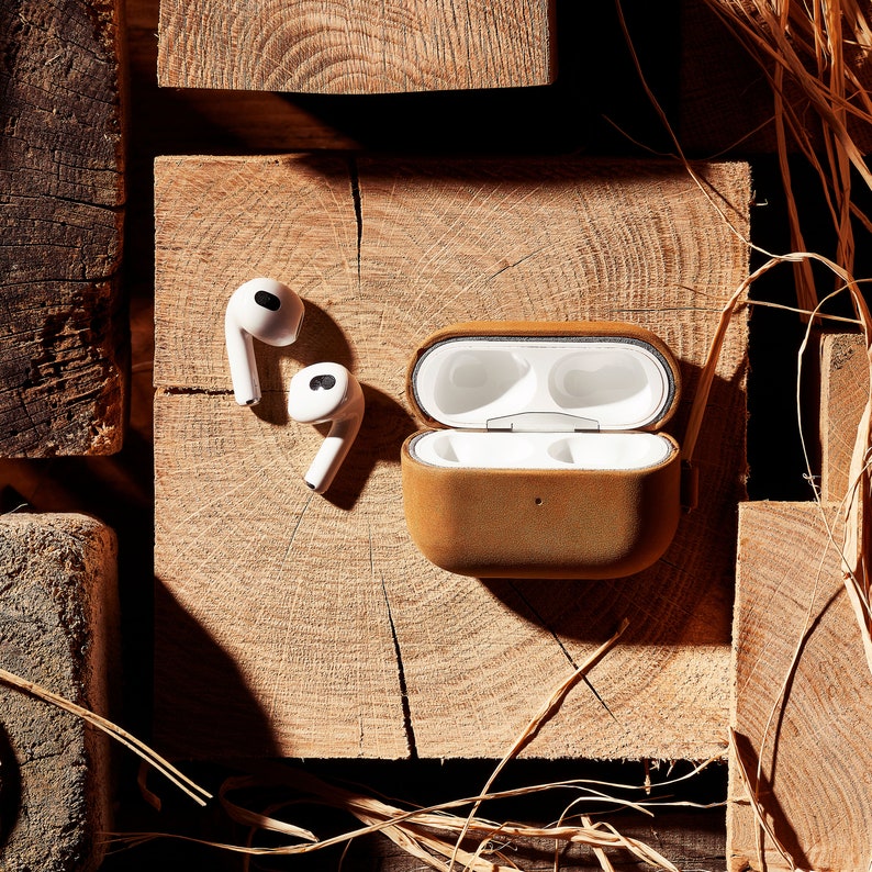 May include: A brown leather case for wireless earbuds with two white earbuds sitting next to it on a wooden surface.