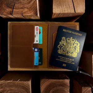 Pu&ograve; includere: Porta passaporto in pelle marrone aperto, con fessure per carte e un passaporto britannico. Il porta passaporto ha un bordo strutturato di colore chiaro. Le carte di credito sono visibili nelle fessure. Il passaporto &egrave; blu con scritte dorate.