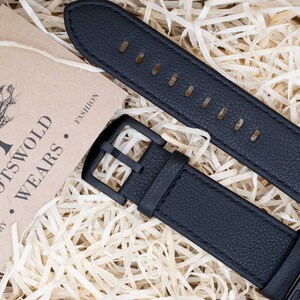 May include: A black textured watch strap with a matching black buckle and connector, accompanied by a tan card featuring a tree illustration and the printed text COTSWOLD WEARS COUNTRY FASHION, displayed on a bed of light-colored wood shavings.
