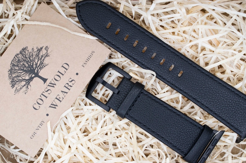 May include: A black textured watch strap with a matching black buckle and connector, accompanied by a tan card featuring a tree illustration and the printed text COTSWOLD WEARS COUNTRY FASHION, displayed on a bed of light-colored wood shavings.