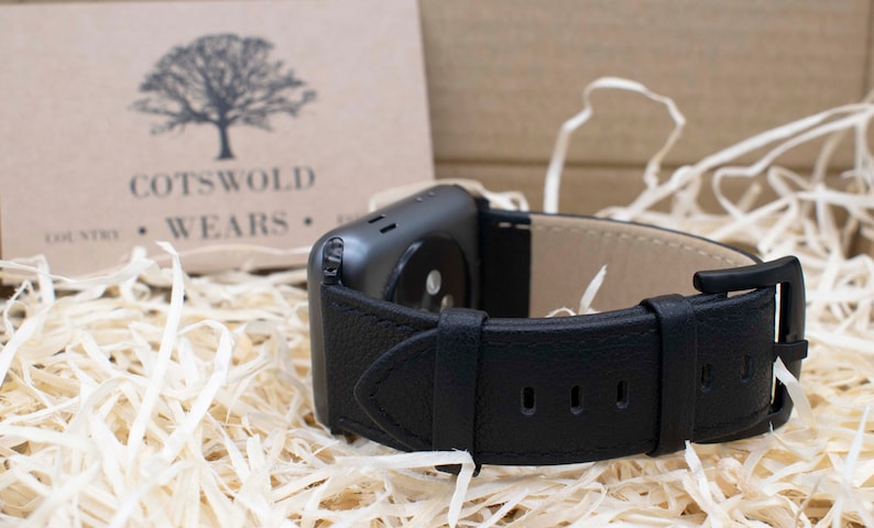 May include: A black leather watch strap attached to a dark gray smartwatch casing. The strap features a textured exterior, a smooth beige interior lining, and a black metal buckle. A tan card with a tree logo and the text COTSWOLD COUNTRY WEARS is visible.