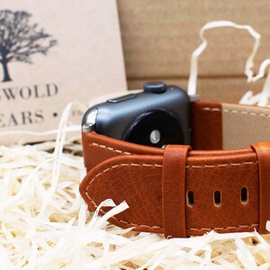 Pu&ograve; includere: Un cinturino per orologio in pelle marrone con cuciture visibili e una fibbia in metallo argentato fissato a uno smartwatch grigio scuro. Sullo sfondo, un cartoncino beige mostra il logo di un albero e la scritta COTSWOLD COUNTRY WEARS.