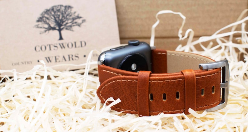 Pu&ograve; includere: Un cinturino per orologio in pelle marrone con cuciture visibili e una fibbia in metallo argentato fissato a uno smartwatch grigio scuro. Sullo sfondo, un cartoncino beige mostra il logo di un albero e la scritta COTSWOLD COUNTRY WEARS.