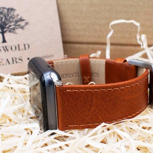 Pu&ograve; includere: Uno smartwatch grigio scuro con un cinturino in pelle marrone chiaro con cuciture a vista e una fibbia in metallo argentato. Il cinturino reca la scritta GENUINE LEATHER. &Egrave; visibile un cartoncino beige con logo ad albero nero e testo COTSWOLD WEARS.