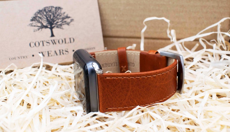 Pu&ograve; includere: Uno smartwatch grigio scuro con un cinturino in pelle marrone chiaro con cuciture a vista e una fibbia in metallo argentato. Il cinturino reca la scritta GENUINE LEATHER. &Egrave; visibile un cartoncino beige con logo ad albero nero e testo COTSWOLD WEARS.