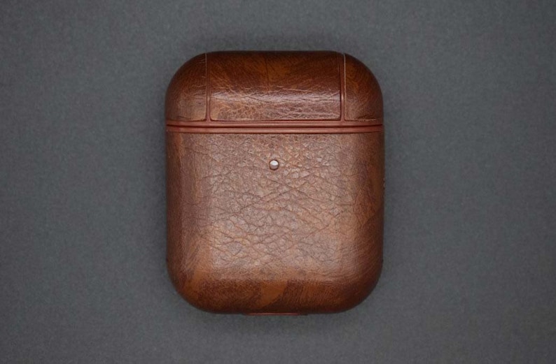 May include: A brown leather case for wireless earbuds. The case has a textured surface and a red trim around the edges.