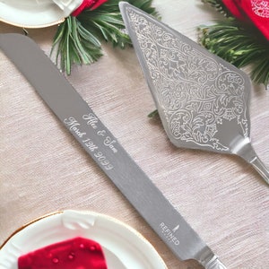 May include: A silver cake knife and server set with an ornate design. The knife has the text "Alex & Sam March 12th 2023" engraved on the blade. The server has a decorative floral pattern. The set is on a white background with red accents.