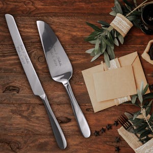 May include: A silver cake knife and server set with engraved text, "James & Natalie, October 5, 2024". The set is on a wooden surface with a brown envelope and green foliage.