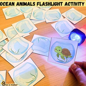 May include: Ocean Animals Flashlight Activity set featuring multiple square cards with illustrations of empty fish bowls and one card revealing a green sea turtle inside a bowl when illuminated by a blue-tinted flashlight beam.