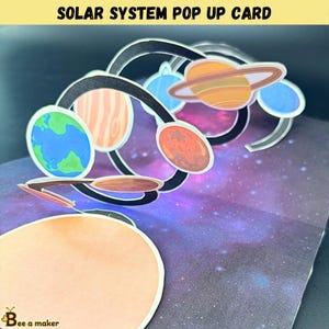 Puede incluir: Una tarjeta desplegable con un diseño colorido del sistema solar. La tarjeta muestra planetas como la Tierra, Marte y Saturno, con un fondo de galaxia. El texto "SOLAR SYSTEM POP UP CARD" está en la parte superior.