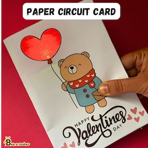 May include: A white card with a red heart balloon and a brown bear holding a red and white scarf. The bear is wearing a blue jacket and holding a small brown envelope. The card says "Happy Valentines Day" in black cursive writing.