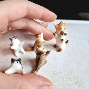 May include: A collection of six miniature ceramic cat figurines in various poses and colors, including white with grey markings, brown tabby, and orange. Each small cat features painted eyes and ears, standing in upright or reaching positions.