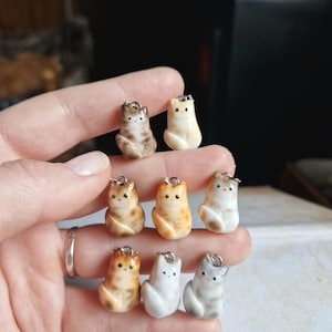 May include: Eight small ceramic cat figurines with different colour patterns. The cats are all sitting with their tails curled around their bodies. They are painted in shades of white, brown, orange, and grey.