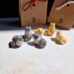 Può includere: Cinque statuine di gatto in ceramica, due sono grigie e tre sono color crema. Le statuine sono sedute su una superficie bianca. Le statuine sono tutte di dimensioni diverse e hanno pose diverse.