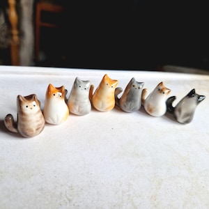 Op de afbeelding: Een rij van acht kleine keramische kattenbeeldjes in verschillende kleuren, waaronder grijs, wit, oranje en bruin. De katten zitten allemaal rechtop met hun staarten opgerold achter zich.