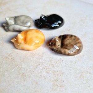 Op de afbeelding: Vier keramische kattenbeeldjes, elk in een andere kleur: grijs, zwart, oranje en bruin. De katten liggen opgerold in een slaaphouding.