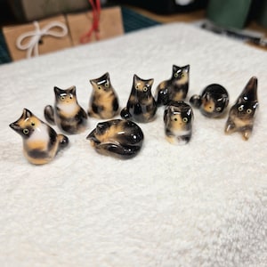 以下が含まれることがあります： 様々なポーズをとった9体の小さな陶器製の猫の置物。各猫は黒、茶、白の斑点がある三毛猫模様が特徴です。座っている姿、横になっている姿、伸びをしている姿などがあり、すべてに目が描かれています。
