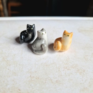 Può includere: Tre statuette di gatto in ceramica, nere, grigie e arancioni. Ogni gatto è seduto con la coda arricciata. I gatti hanno occhi verdi o gialli e sono su una superficie chiara.