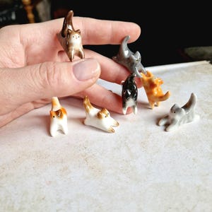 May include: A collection of seven small ceramic cat figurines in various poses. The cats are painted in shades of white, grey, brown, orange, and black.