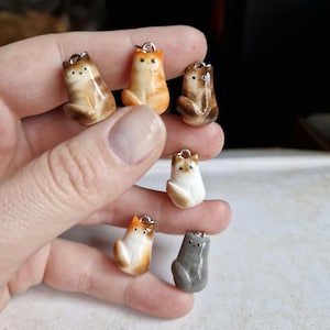 以下が含まれることがあります： 異なる色と模様の小さなセラミック製の猫の置物6個。猫は直立して座っており、シンプルで漫画のようなデザインです。すべて銀色のチャームが付いています。