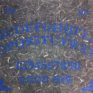 May include: A black and white Ouija board with blue lettering. The board features the alphabet, numbers 1-9 and 0, the words "yes", "no", and "goodbye", and two symbols, a star and a crescent moon.