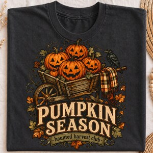 Op de afbeelding: Een antracietgrijs t-shirt met een afbeelding van een houten kruiwagen gevuld met oranje pompoenen, een geruite sjaal en een zwarte kraai. De tekst luidt PUMPKIN SEASON en haunted harvest club, omringd door herfstbladeren en ranken.