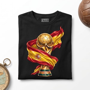 Może przedstawiać: Czarny t-shirt z nadrukiem graficznym przedstawiającym złote trofeum piłkarskie owinięte powiewającą hiszpańską flagą. Wzór zawiera czerwone i żółte barwy flagi z widocznym godłem narodowym. Koszulka posiada okrągły dekolt.