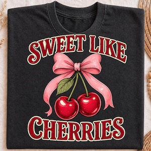 May include: A black crew neck t-shirt featuring a graphic of two bright red cherries with a green leaf and a large light pink bow. The text SWEET LIKE CHERRIES is printed in a bold, dark red font with a white outline above and below the illustration.