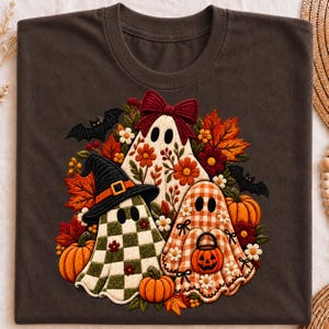Può includere: Una t-shirt marrone scuro con una grafica centrale di tre fantasmi. Un fantasma indossa un cappello da strega, uno ha un fiocco rosso e uno tiene in mano un secchiello a forma di zucca. Sono circondati da zucche arancioni, foglie e pipistrelli.