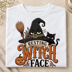 May include: A white t-shirt featuring a graphic of a black witch hat, a broom, a black cat, and an orange pumpkin. The text "RESTING WITCH FACE" is printed in large orange and black lettering. The design includes small floral accents.