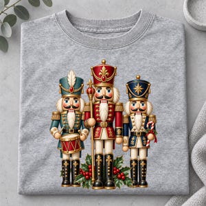 May include: A heather grey t-shirt featuring a graphic of three nutcrackers. The left nutcracker wears teal, the centre wears red, and the right wears blue. They are decorated with gold accents and stand above green holly leaves with red berries.