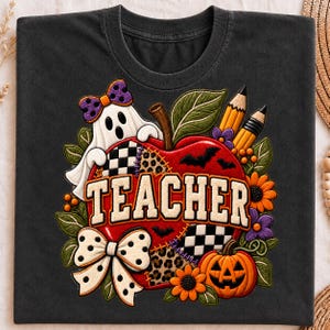 May include: A charcoal gray t-shirt featuring a graphic of a red apple with the word TEACHER in white letters. The design includes a white ghost with a purple bow, two yellow pencils, a small orange pumpkin, sunflowers, and black bats.