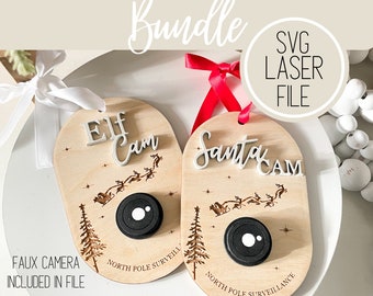 Op de afbeelding: Twee houten kerstversieringen met een nep-cameralenzen. De versieringen zijn versierd met de woorden "Elf Cam" en "Santa Cam" en de tekst "Toezicht Noordpool".