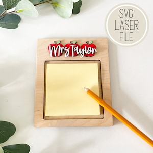 May include: Wooden note holder with a cutout for a sticky note. The holder has three red apple cutouts with the text "Mrs. Taylor" above the note area. A yellow pencil is lying on the note.