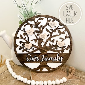 Op de afbeelding: Een ronde houten familieteken met een donkerbruine afwerking en witte achtergrond. Het boomontwerp heeft hartvormige naamlabels. Het bord leest "Our Family" in wit script. De woorden "SVG LASER FILE" staan in een cirkel rechtsboven.