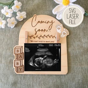 Puede incluir: Cartel de anuncio de embarazo de madera con las palabras "Coming soon!" y "We can't wait to meet you!". El cartel incluye una imagen de ultrasonido y la palabra "BABY" deletreada con bloques de madera. El cartel es un archivo láser.
