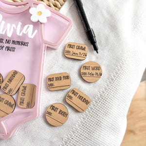 Puede incluir: Una caja de recuerdos rosa con forma de body de beb&eacute; con el nombre "Olivia" y el texto "Primeras veces, Grandes recuerdos de las primeras veces". Incluye fichas de madera para hitos como "Primer gateo" y "Primera palabra". Tambi&eacute;n se ve un rotulador negro.