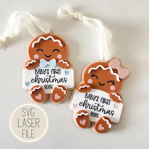 Pode incluir: Dois ornamentos de boneco de gengibre com fitas brancas. Cada figura castanha segura uma placa branca com a inscrição Baby's First Christmas 2026. Um tem uma gravata borboleta azul clara, o outro um laço rosa, com pequenos corações.
