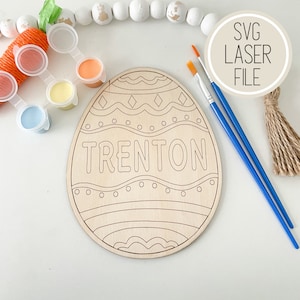 May include: A wooden Easter egg shaped craft with the name TRENTON in the center. The egg is decorated with a wavy line pattern and a diamond pattern. The craft comes with paint pots and paintbrushes.
