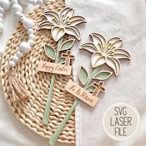 May include: Two wooden Easter lily decorations with the text "Happy Easter" and "He is Risen". The lilies have layered petals in cream and brown, green leaves, and a cross. The stems are painted green.