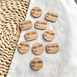 Puede incluir: Un conjunto de marcadores de hitos de madera con texto negro sobre tela blanca. Los marcadores tienen varias formas, incluyendo c&iacute;rculos y rect&aacute;ngulos. Cada marcador tiene una frase como "Primera risa" o "Primer cumplea&ntilde;os" y una l&iacute;nea para la fecha.